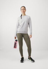 Grå hettegenser, olivengrønne leggings, svarte og gule joggesko, holder en rosa vannflaske med svart "PUMA"-logo.