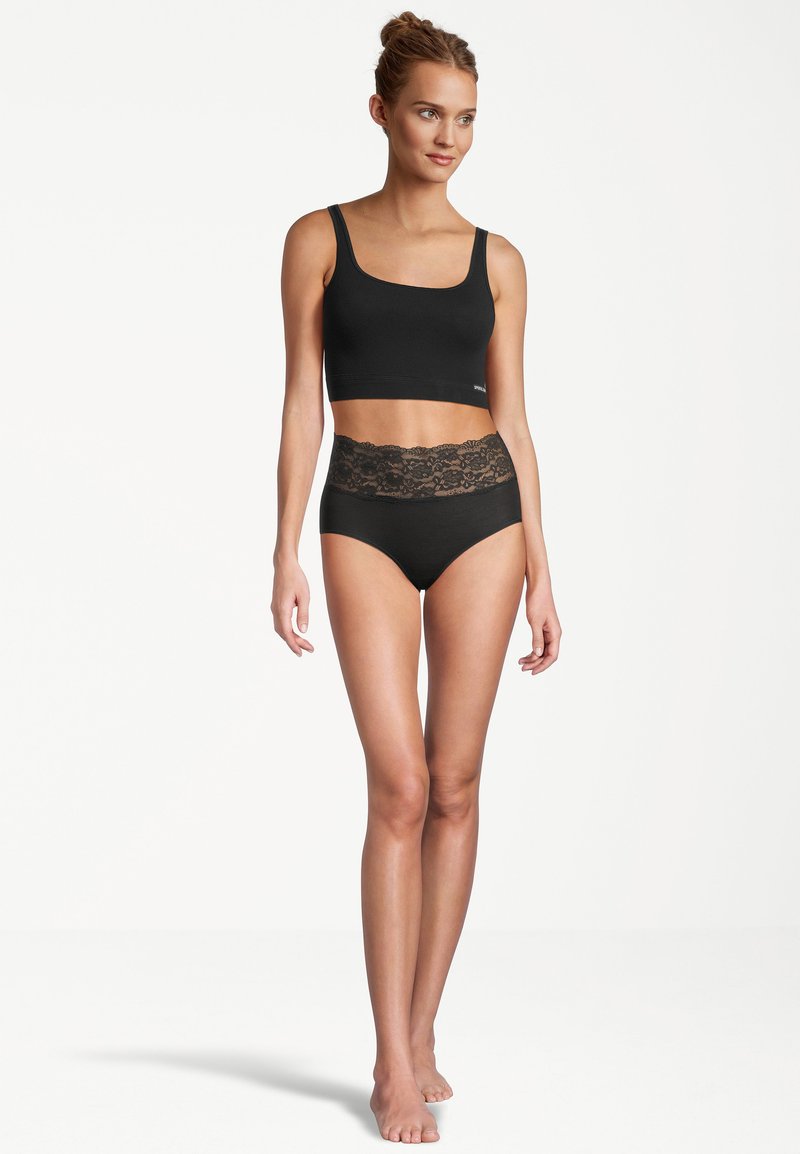 Donna che indossa un top corto nero e un perizoma in pizzo nero a vita alta, in piedi a piedi nudi su uno sfondo bianco, mentre guarda verso destra.