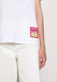 Biała bawełniana koszulka z kolorową naszywką w różowe i żółte akcenty, z napisem "Karl Lagerfeld Paris" wzdłuż dolnej krawędzi.