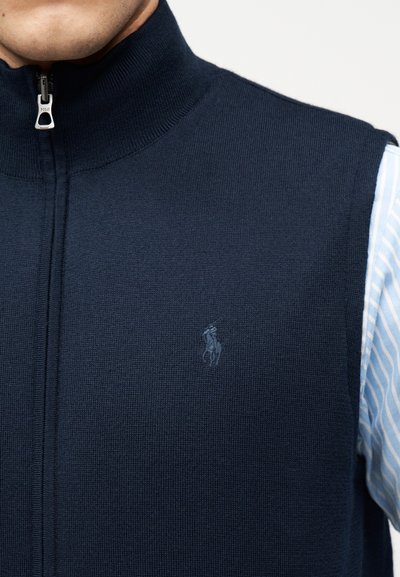 Κοντινή λήψη ενός ατόμου που φοράει μπλε σκουρόχρωμο πλεκτό γιλέκο με φερμουάρ πάνω από ένα ανοιχτό μπλε και λευκό ριγέ πουκάμισο, εμφανίζοντας το γιακά και το φερμουάρ του γιλέκου.