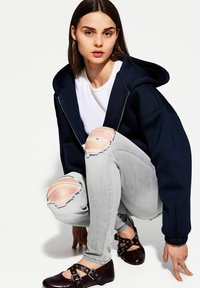 Sweat-shirt zippé navy, t-shirt blanc, jeans gris déchirés avec des genoux troués, et chaussures marron foncé avec des boucles. Modèle accroupi sur fond blanc.