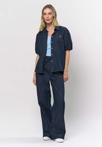 Donker denim, overhemd met knopen en pofmouwen, gedragen over een lichtblauwe tanktop. Bijpassende donkere denim broek, witte sneakers en eenvoudig ontwerp.