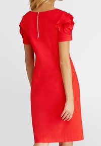 Robe rouge avec manches courtes volantées et une fermeture éclair discrète au dos. Tissu lisse, coupe ajustée, longueur genou, couleur unie.