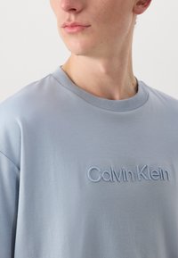 Light blue cotton t-shirt with a round neck, featuring embroidered "Calvin Klein" text on the front. Soft texture, relaxed fit.