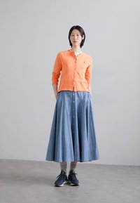 Orange button-up cardigan over a white shirt, paired with a long light blue denim skirt and dark sneakers. Smooth textures and relaxed fit.