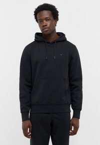 Mężczyzna w czarnej bluzie z kapturem, z przednią kieszenią i sznurkami, stoi na jednolitym białym tle, zwrócony przodem.