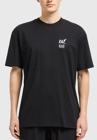 Man i svart oversized t-shirt med liten vit EA7-logga på vänster bröst, stående mot en enkel ljusgrå bakgrund.