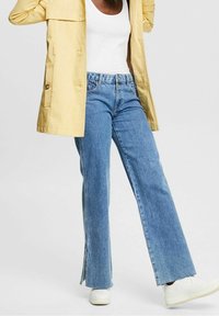 Veste légère jaune clair portée sur un débardeur blanc, associée à un jean flare bleu avec des fentes latérales et des baskets blanches.