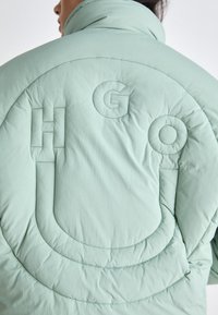 Giacca gonfiabile verde menta con colletto alto, caratterizzata da ricami con "HGO" e motivi di cucitura curvi sul retro. Materiale testurizzato e leggero.