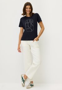 Marinblå bomullströja med vit grafisk text fram, kombinerad med krämfärgade vida byxor och sneakers med gröna accentfärger.