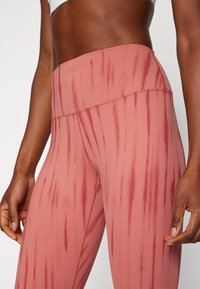 Leggings de cor pêssego, de cintura alta, com riscas verticais em tons mais escuros, feitos de um tecido suave e elástico. Detalhes de costura são visíveis.