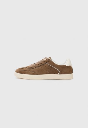 Lage bruine suède sneaker met beige zool, beige veters, wit hielaccent en perforatiedetails aan de zijkant.