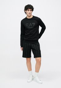 Fiatal férfi fekete EA7 Emporio Armani pulóverben és hozzá illő rövidnadrágban, fehér zokniban és fehér tornacipőben fehér háttér előtt áll.