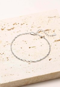 Pulsera de alambre retorcido de plata con un pequeño acento de perla, que presenta un cierre de langosta y una cadena de extensión sobre un fondo texturizado en tonos neutros.