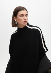 Žena s krátkými hnědými vlasy, oblečená v nadměrném černém svetr s bílými pruhy na ramenech a velkými stříbrnými náušnicemi, hledící do strany.