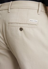 Beige cotton trousers with a back pocket featuring a round brown button and a stitched label that reads "Marc O'Polo." Smooth texture.