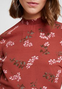 Kvinde iført en rust-rød blomsterbluse med pink blomster og grønne blade, med en rynket høj hals og guld rektangulære øreringe.