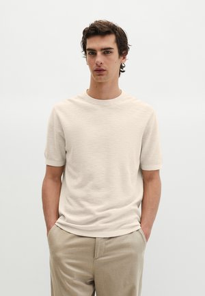 Młody mężczyzna w kremowym krótkim swetrze z rękami w kieszeniach beżowych spodni na jasnym jednolitym tle.