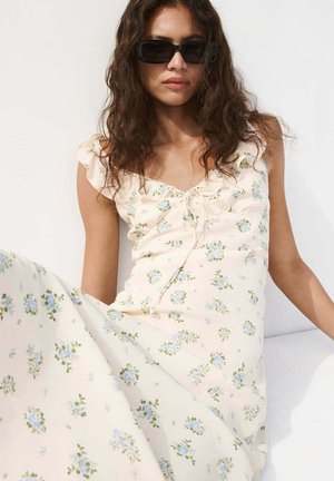 Kvinde iført rektangulære sorte solbriller og en cremefarvet blomsterkjole med blå blomster, siddende mod en hvid baggrund.