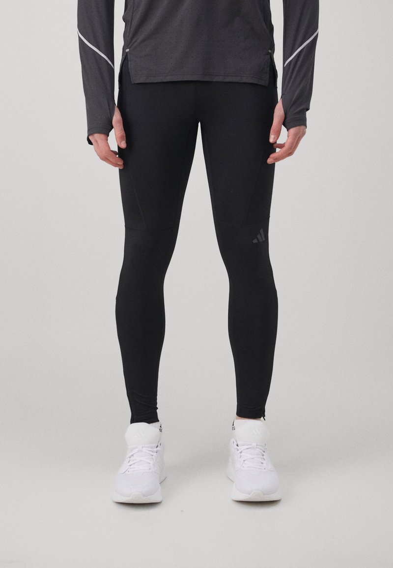 Leggings de sport noirs en matériau extensible, avec un design ajusté et une marque discrète. Portés avec des chaussures de sport blanches.