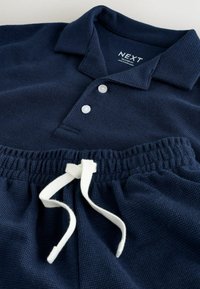 Polo bleu marine texturé avec deux boutons blancs associé à un pantalon à taille élastique bleu marine avec cordon blanc.