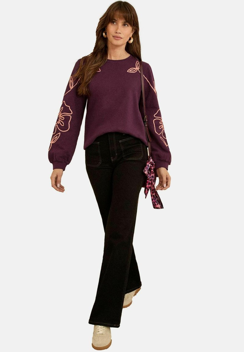 Sweat-shirt violet avec broderie florale sur les manches, associé à un jean noir évasé et des baskets beiges. Comprend un accessoire de sac à motifs.