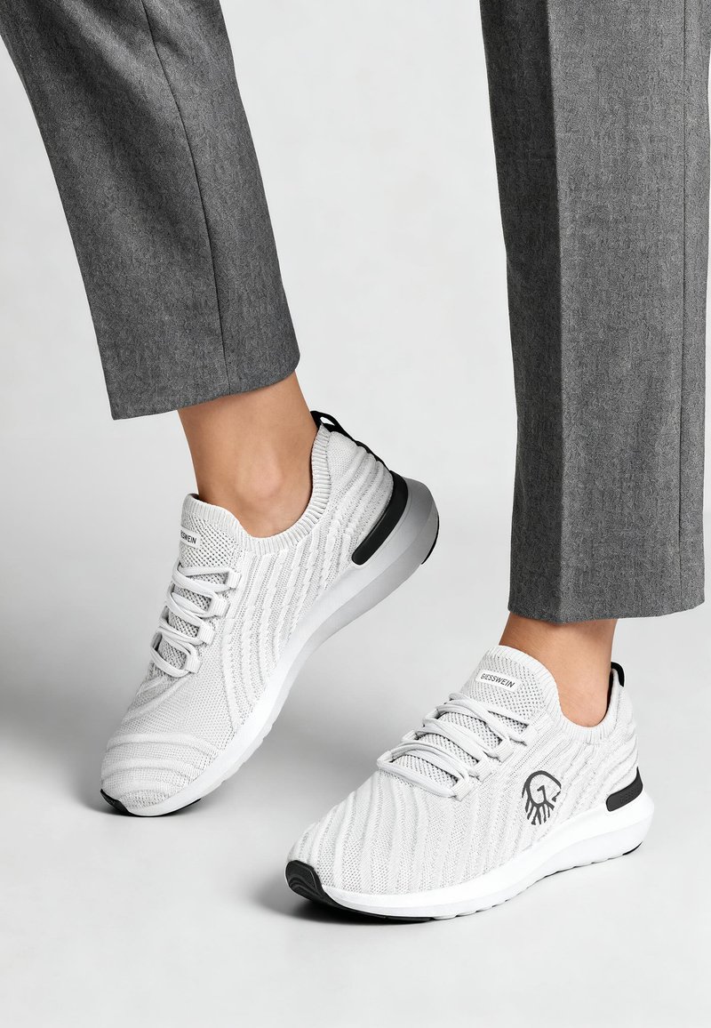 Personne portant un pantalon gris et des baskets blanches en maille avec des accents noirs, se tenant sur une surface gris clair.