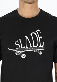 Sort t-shirt med et hvidt grafisk design, der viser "SLADE" over et skateboard-design med detaljerede hjul og brædemodel.