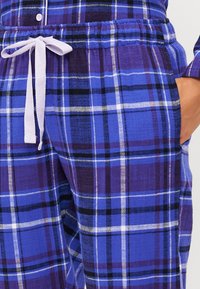 Pantalons de pyjama en tartan violet en tissu doux. Dotés d'une taille élastique avec un lien violet clair, de poches latérales et de lignes blanches en damier.