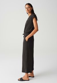 Jumpsuit noir sans manches avec une finition texturée, taille avec cordon de serrage, poches latérales et jambes larges. Assorti à des sandales à glissière noires simples.
