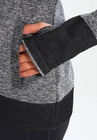 Grå og sort athletic top med et tekstureret mønster, der har en sidelomme og tommelfinger-ærmegab, lavet af et strækbart, åndbart materiale.