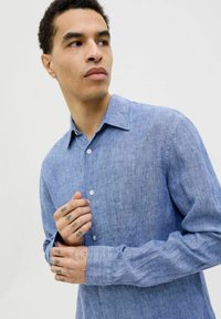 Camisa de lino azul claro con cuello estándar, mangas largas y cierre de botones. Textura visible y detalle de patrón de tejido sutil.
