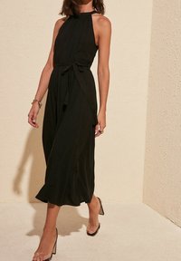 Zwarte halternekjurk met een vloeiende uitstraling, strik taille en lengte tot halverwege het scheenbeen. Model draagt doorzichtige sandalen met hakken. Gladde stoftextuur.