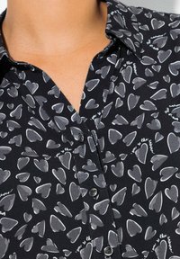 Schwarze Bluse mit einem Kragen, der ein sich wiederholendes Herzmuster in Grautönen zeigt. Enthält Knöpfe an der Vorderseite zum Verschließen.