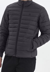 Giacca imbottita nera con collo alto, caratterizzata da cuciture orizzontali, zip frontale e tasche laterali. Texture liscia e design minimalista.