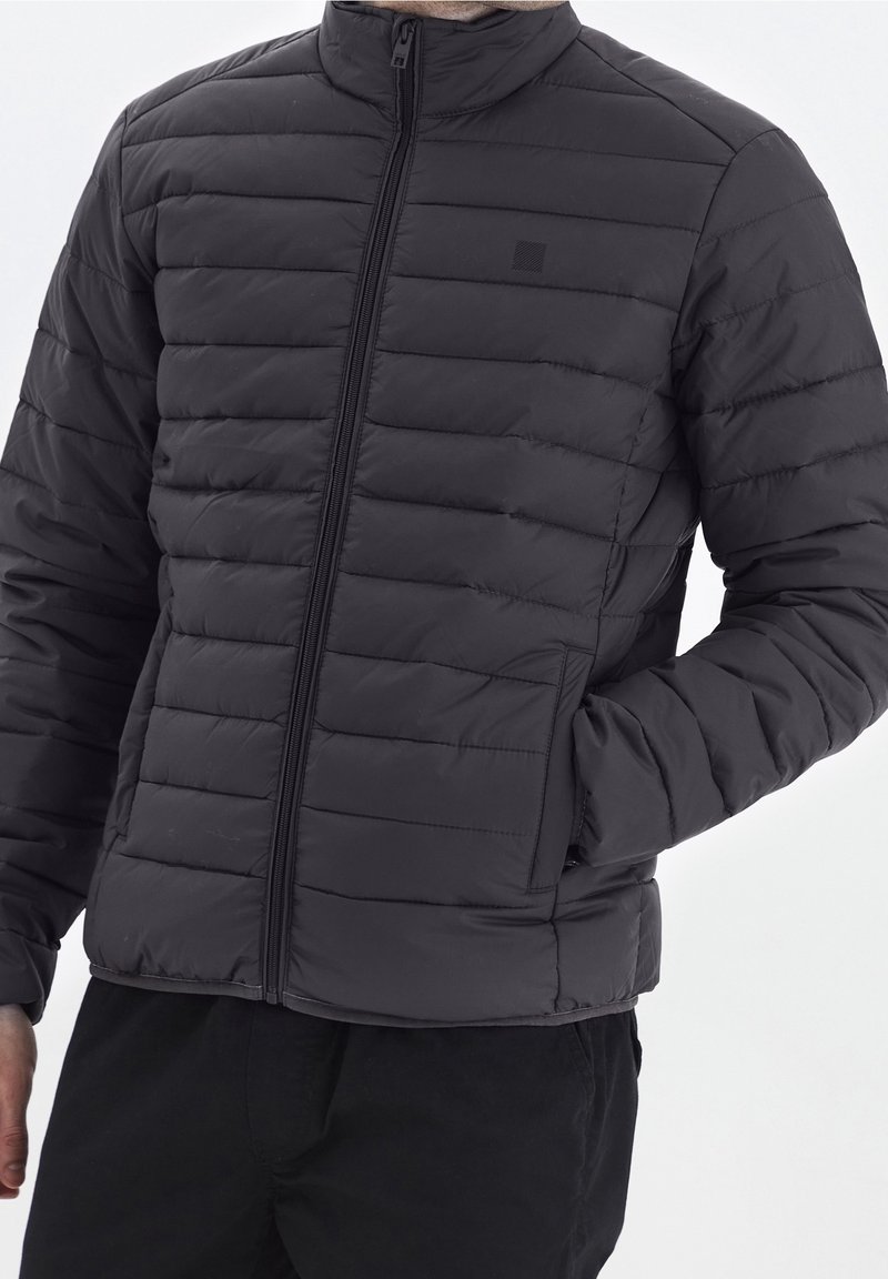 Giacca imbottita nera con collo alto, caratterizzata da cuciture orizzontali, zip frontale e tasche laterali. Texture liscia e design minimalista.