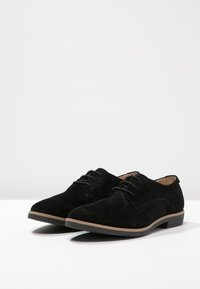 Scarpe in suede nere con una finitura liscia, punta rotonda e lacci. Presentano una suola beige in contrasto e un sottile dettaglio di cucitura sui lati.
