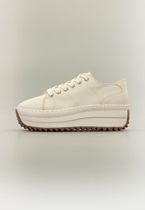 Crèmekleurige platformsneaker met dikke witte zool en bruin zigzagprofiel, vetersluiting en bovenwerk van canvas.