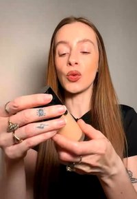 Kvinne med langt rødt hår holder og undersøker en foundationflaske, iført svart topp og ringer, mot en ensfarget bakgrunn.