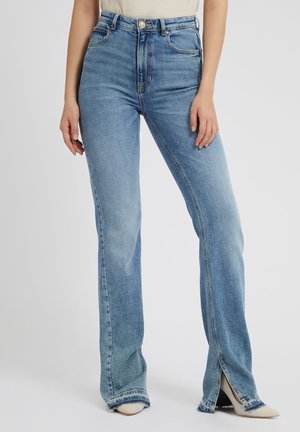 Vrouw draagt lichtblauwe jeans met hoge taille en vooraan gespleten pijpen bij de enkels, gecombineerd met witte schoenen met puntige neus en een beige top ingestopt.