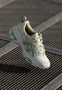 Baskets de sport en cuir beige clair avec des accents verts, présentant une semelle épaisse et un dessus texturé. Lacets et languette au talon inclus.