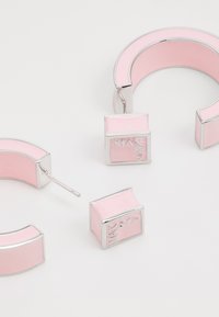 Boucles d'oreilles roses avec une finition brillante et des accents argentés. Comprend deux formes géométriques : des anneaux incurvés et des clous carrés avec un logo.