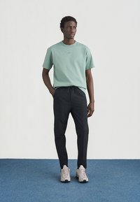 Hellgrünes Baumwoll-T-Shirt mit kleinem Logo, kombiniert mit schwarzer Hose und beigen Sneakers, stehend auf einem blauen Teppich vor einem schlichten Hintergrund.