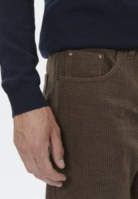 Ruskeat samettihousut, joissa on pystyribbitys, napit ja sivutaskut. Yhdistetty tummansiniseen neulepaitaan.