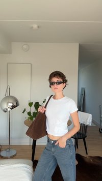 Valkoinen lyhyt t-paita, korkeavyötäröiset siniset farkut, ruskea mokkabag. Sisustus, jossa on hopeinen lattiavalaisin, peili ja vihreä kasvi.