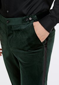 Pantaloni verdi in velluto con una texture liscia, caratterizzati da una parte anteriore piatta e tasche laterali. Strisce laterali nere e dettagli con bottoni in vita.