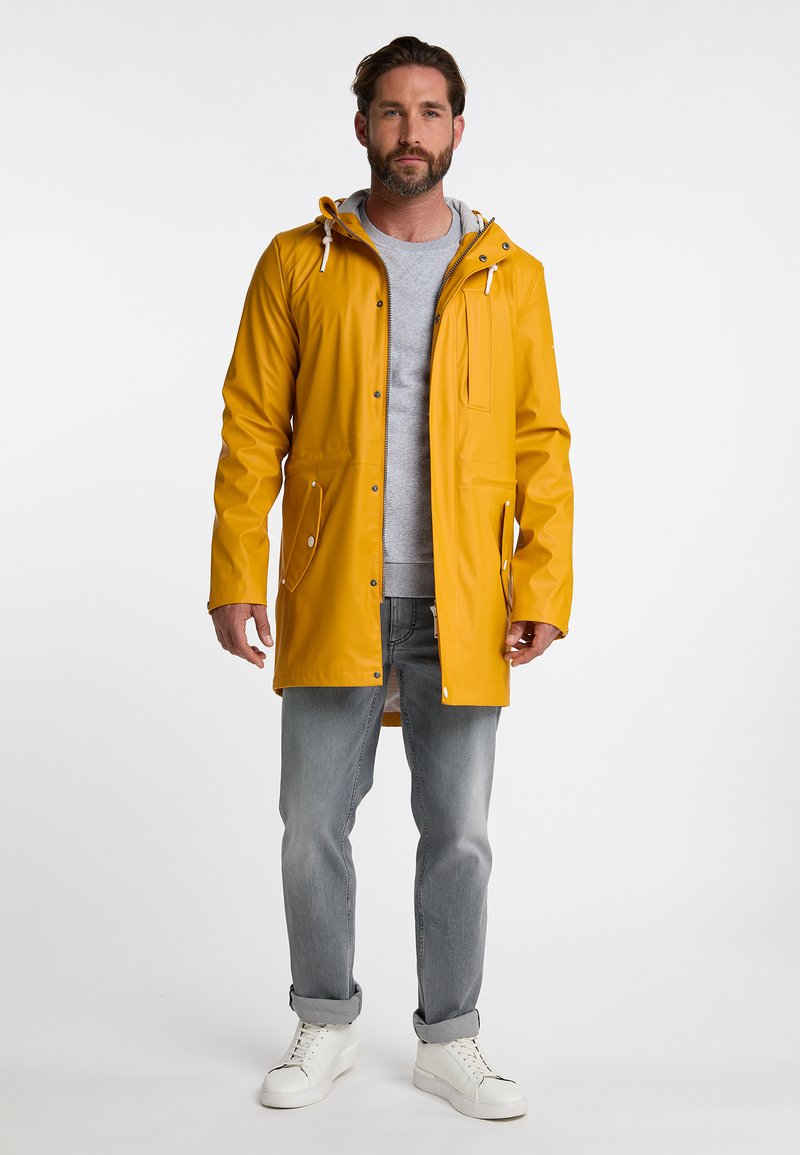 Geel waterdichte jas met een capuchon, ritssluiting aan de voorkant, twee zijzakken en trekkoorddetails, gedragen over een grijze sweatshirt en spijkerbroek.