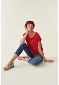 Top corto rojo de manga corta con un ajuste holgado, vaqueros cortos azules y mocasines de ante beige con un acento dorado, sentada contra un fondo neutro.