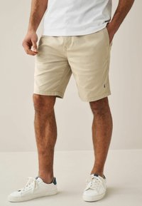 Bermuda beige con un design fronte piatto, passanti per cintura e tasche laterali. Abbinati a sneaker bianche con accenti blu.