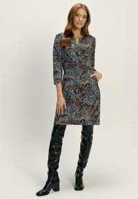 Bunt gemustertes Kleid mit Dreiviertelärmeln, einem Ausschnitt mit Schlüsselöffnung und seitlichen Taschen, kombiniert mit langen, glänzenden, kniehohen schwarzen Stiefeln.